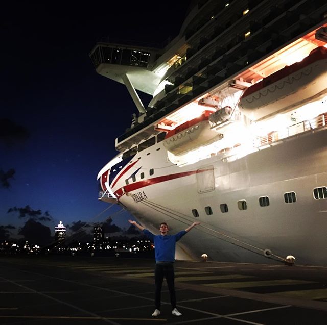 Adam with arms in air at night, large cruise ship and cityscape in background